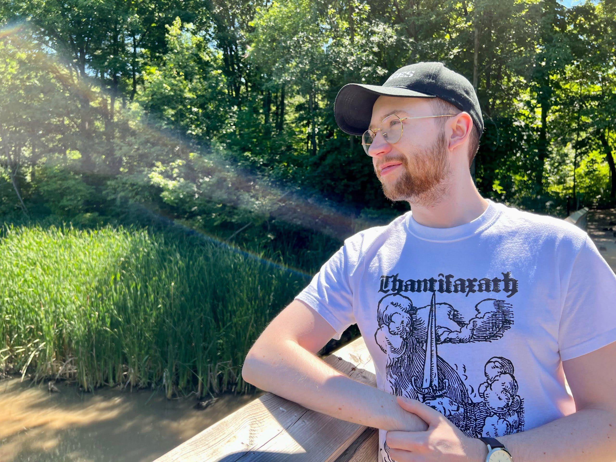 Rory Hoy outside and suprisingly not on the computer, on a bridge overlooking a swampy marsh. There is a streak of chromatic aberration from the sunlight across the image.